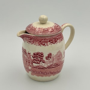 May include: A cream-colored ceramic teapot with a lid, featuring a detailed red transferware pattern of trees, a fence, and architectural structures. The teapot has a curved handle, a small spout, and a round knob on the center of the lid.