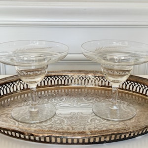 May include: Two clear margarita glasses with etched leaf designs sit on a silver-plated serving tray. The tray has a decorative pierced edge and a detailed floral pattern in the center. The glasses are centered on the tray, ready for serving.