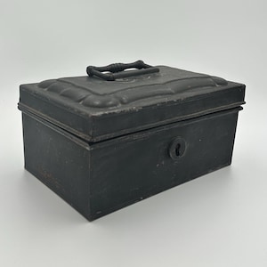 May include: A rectangular black metal lockbox featuring a hinged lid with a raised decorative border and a central handle. The front panel includes a keyhole mechanism. The surface shows signs of wear and a matte finish throughout the exterior.