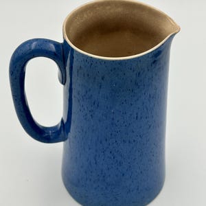 May include: A ceramic pitcher featuring a vibrant blue exterior with a speckled finish and a curved handle. The interior of the vessel is a smooth, light beige color, and the rim includes a small, integrated pouring spout for liquid dispensing.