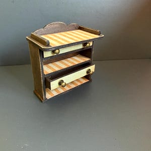 Puede incluir: Una pequeña cómoda de madera con un marco marrón oscuro y dos cajones extraíbles con rayas verticales de color crema y naranja claro. La unidad tiene un borde superior festoneado y pequeños pomos redondos de metal color latón en cada cajón.