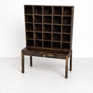Puede incluir: Un mueble de madera marrón oscuro con múltiples compartimentos cuadrados pequeños. El mueble se asienta sobre una mesa con un solo cajón y un pomo de latón. La pieza tiene una estética vintage y está sobre un fondo blanco.