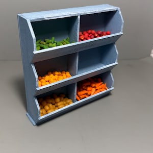May include: A miniature, light blue wooden produce stand with six compartments. Each compartment holds a different type of colorful produce, including green beans, red berries, orange and yellow fruits, and carrots. The word "Vegetables" is written on the stand.
