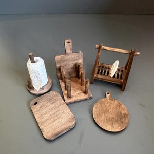 May include: A collection of miniature wooden kitchen accessories including a paper towel holder with a white roll, a rectangular cutting board, a circular cutting board, a vertical utensil holder, and a small dish drying rack with one white plate.