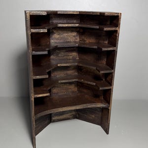 May include: A wooden corner shelf unit with multiple shelves. The unit is stained in a dark brown color with a slightly distressed finish. The shelves are arranged in a stepped pattern, creating a tiered display space. The unit is designed to fit into a corner.