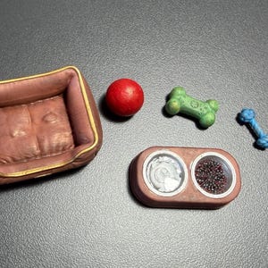 May include: A miniature pet bed, red ball, green bone-shaped toy, blue rope toy, and a double bowl set. The pet bed is brown with a gold trim. The bowls are brown with silver and dark brown contents.