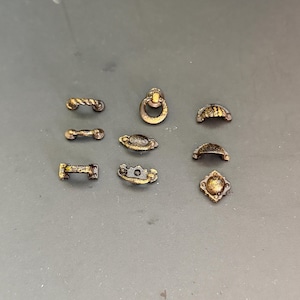 Könnte beinhalten: Eine Sammlung von acht Miniatur-Schubladengriffen und Schrankgriffen aus Metall in dunkler Bronzeoptik mit goldfarbenen Akzenten. Das Set enthält verschiedene Formen wie Ringgriffe, Stangengriffe und Muschelgriffe mit detaillierten Verzierungen.