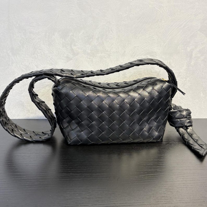 May include: Black woven leather shoulder bag with a zip closure. The bag features a braided strap and a knotted detail on one side. The bag is sitting on a black surface.