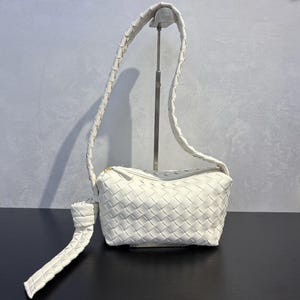May include: A white woven leather crossbody bag with a long, braided strap. The bag has a rectangular shape with a zipper closure and a knotted detail on the strap. The bag is displayed on a black surface.