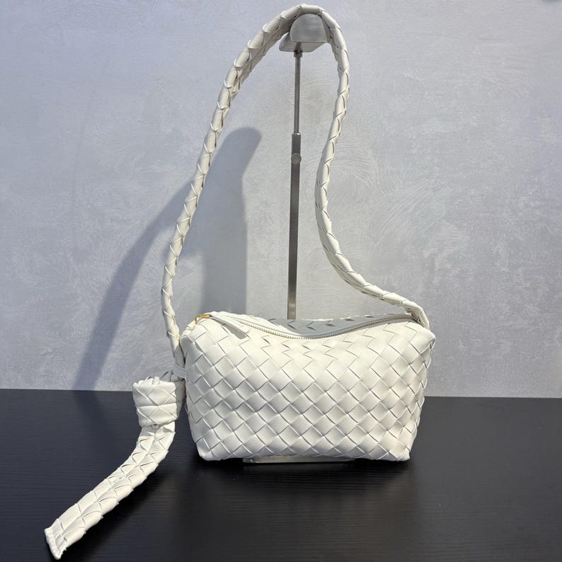 May include: A white woven leather crossbody bag with a long, braided strap. The bag has a rectangular shape with a zipper closure and a knotted detail on the strap. The bag is displayed on a black surface.