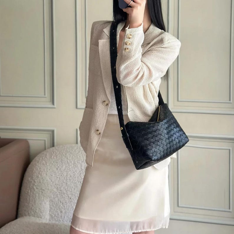 May include: A black woven leather crossbody bag with a long, adjustable strap. The bag is square-shaped and features a zipper closure. The strap is black and appears to be made of a woven material. The bag is worn over a cream-colored outfit.