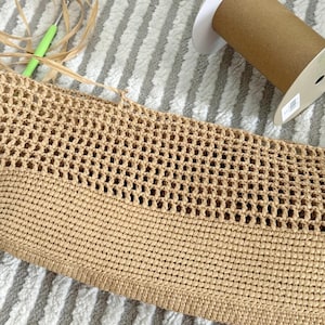 May include: A close-up of a beige crocheted bag with a woven pattern. The bag is made of a natural fiber and has a textured surface.