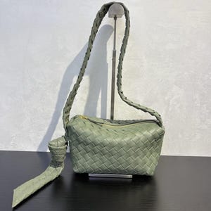 May include: Sage green woven leather shoulder bag with a braided strap and a gold-tone zipper. The bag features a rectangular shape with a knotted detail on the strap. The bag is displayed on a black surface.
