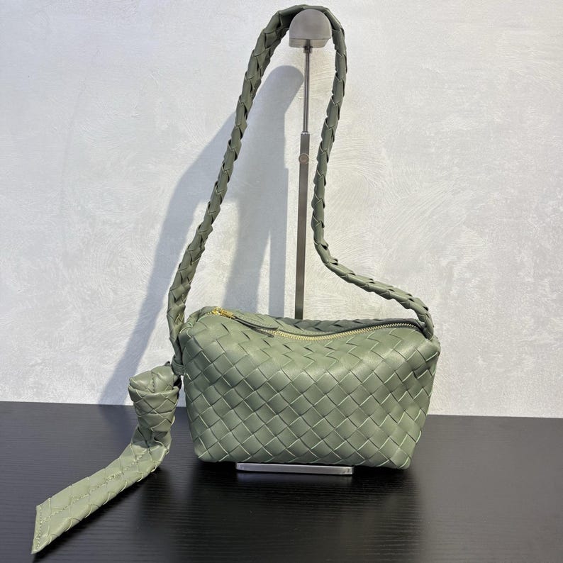 May include: Sage green woven leather shoulder bag with a braided strap and a gold-tone zipper. The bag features a rectangular shape with a knotted detail on the strap. The bag is displayed on a black surface.