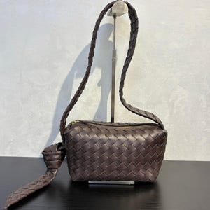 May include: A brown woven leather crossbody bag with a long, braided strap. The bag has a rectangular shape with a zippered top and a knotted detail on the strap. The bag is displayed on a black surface.