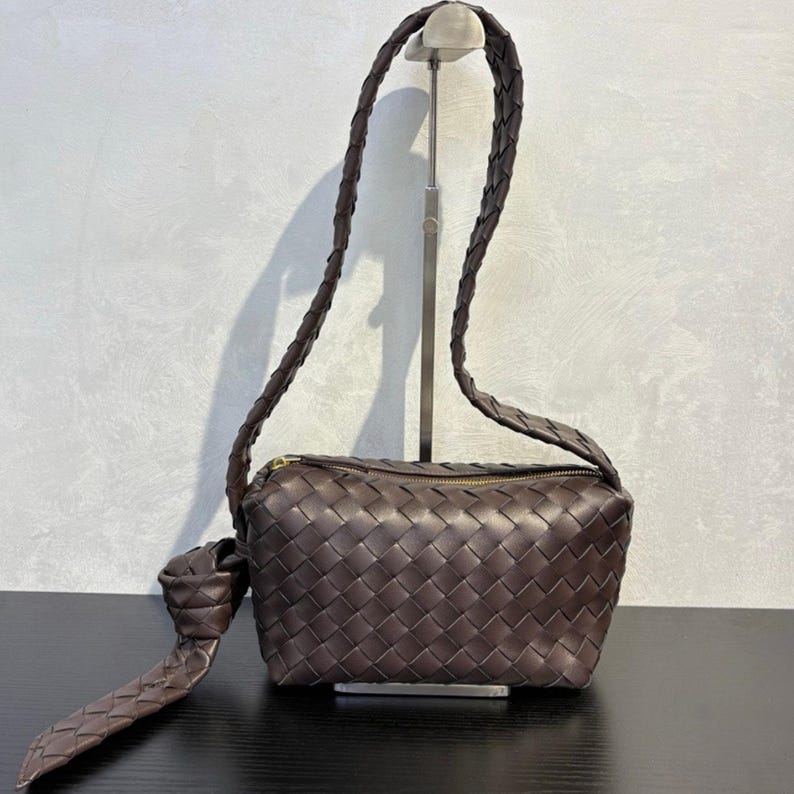 May include: A brown woven leather crossbody bag with a long, braided strap. The bag has a rectangular shape with a zippered top and a knotted detail on the strap. The bag is displayed on a black surface.