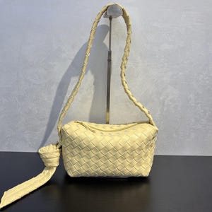 May include: A pale yellow woven leather handbag with a braided shoulder strap. The rectangular bag features a top zipper closure and a knotted detail on one side. The bag is displayed on a black surface.