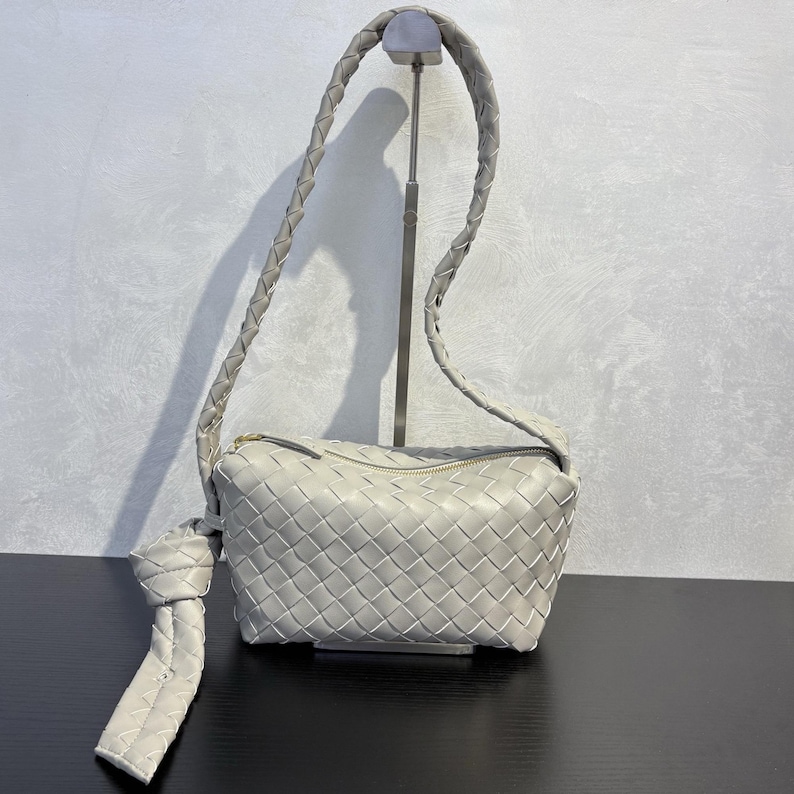 May include: A light gray woven leather handbag with a braided shoulder strap. The bag has a rectangular shape with a zipper closure and a knotted detail on the side. The bag is displayed on a black surface.