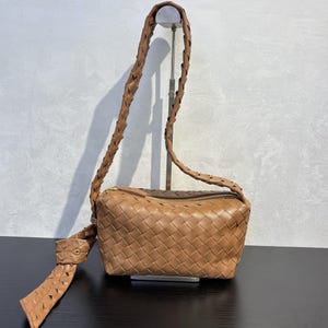 May include: A tan leather crossbody bag with a woven design. The bag has a braided strap and a zipper closure. The strap is knotted at the bottom. The bag is displayed on a black surface.