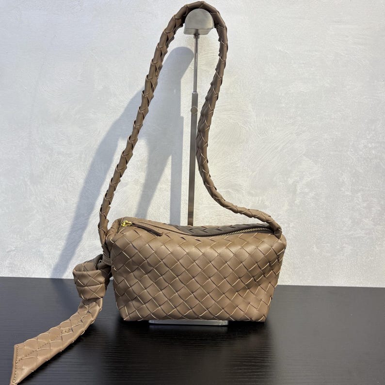 May include: A taupe-colored crossbody bag with a woven design. The bag features a braided strap and a knotted detail on the side. It has a zippered top and a rectangular shape. The bag is displayed on a black surface.