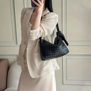 May include: Black woven leather shoulder bag with a knotted strap. The bag has a triangular shape and a zipper closure. The bag is being held by a person wearing a cream-colored blazer and skirt.
