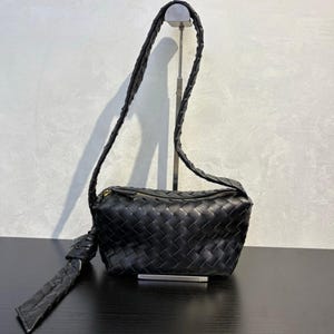 May include: Black woven leather crossbody bag with a long, braided strap. The bag has a rectangular shape with a zipper closure and a knotted detail on one side. The bag is displayed on a black surface.