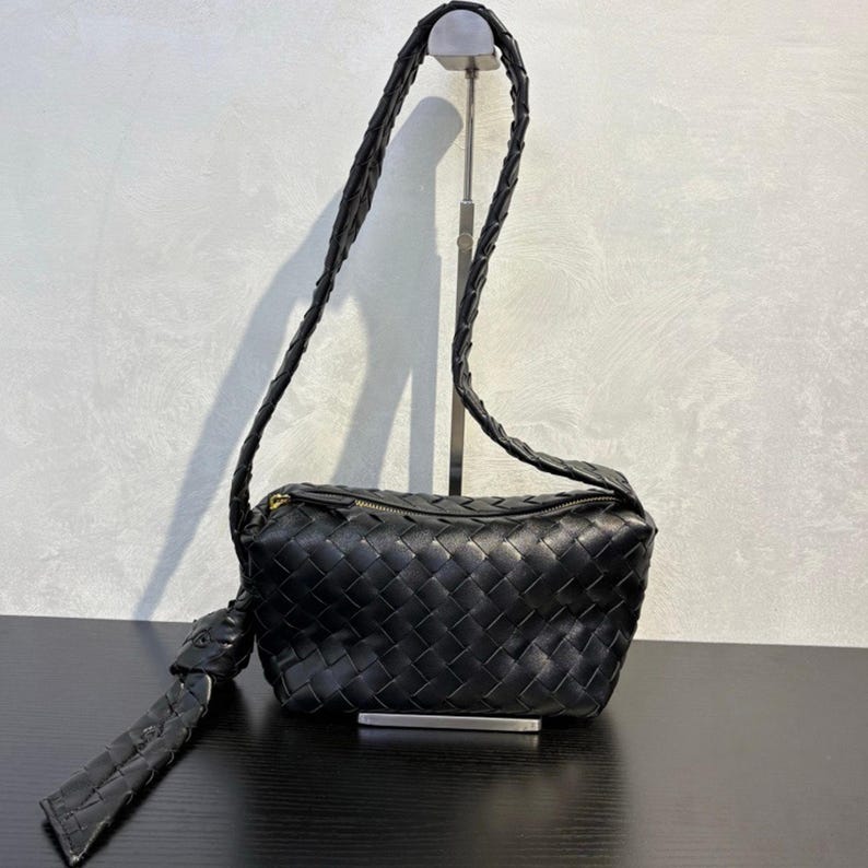 May include: Black woven leather crossbody bag with a long, braided strap. The bag has a rectangular shape with a zipper closure and a knotted detail on one side. The bag is displayed on a black surface.