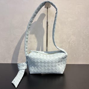 May include: A light blue woven leather shoulder bag with a braided strap and a gold zipper. The bag is displayed on a black surface against a neutral background. The bag's strap is looped over a metal stand.