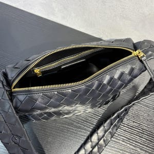 May include: Black woven leather shoulder bag with a gold zipper. The bag has a braided strap and an open interior. The zipper is gold-colored, and the bag is made of a woven leather material.