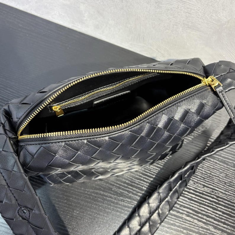 May include: Black woven leather shoulder bag with a gold zipper. The bag has a braided strap and an open interior. The zipper is gold-colored, and the bag is made of a woven leather material.