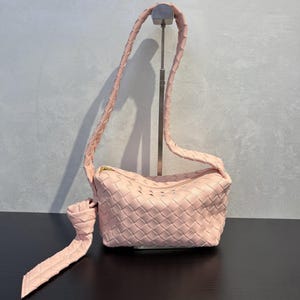 May include: A light pink woven leather shoulder bag with a braided strap and a gold zipper. The bag is displayed on a black surface against a gray background. The strap is long enough to be worn over the shoulder.