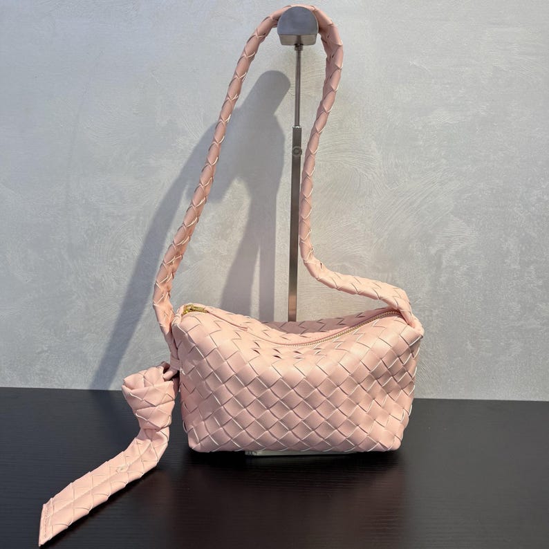 May include: A light pink woven leather shoulder bag with a braided strap and a gold zipper. The bag is displayed on a black surface against a gray background. The strap is long enough to be worn over the shoulder.