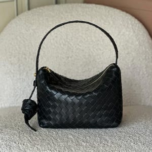 May include: Black woven leather handbag with a curved top and a single handle. The bag features a zipper closure and a decorative leather charm. The bag is a dark, solid color and has a structured shape.