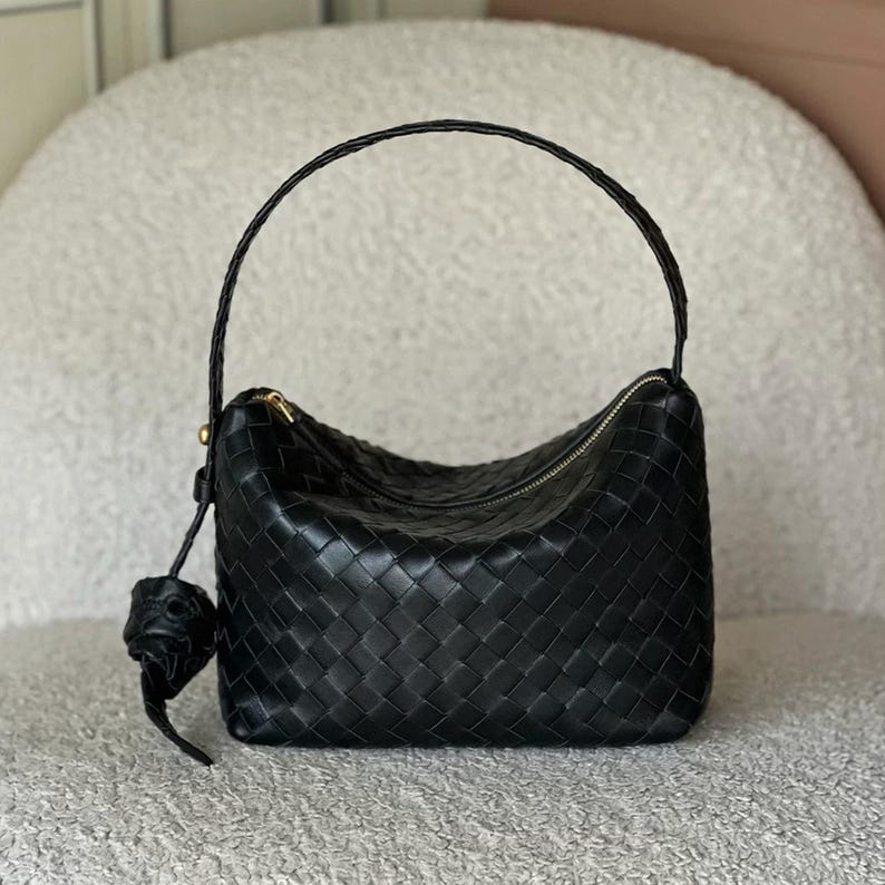 May include: Black woven leather handbag with a curved top and a single handle. The bag features a zipper closure and a decorative leather charm. The bag is a dark, solid color and has a structured shape.