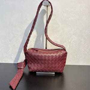May include: A burgundy-colored crossbody bag with a woven design. The bag features a long, braided strap and a zippered top. A decorative knot detail hangs from the side. The bag is displayed on a black surface.