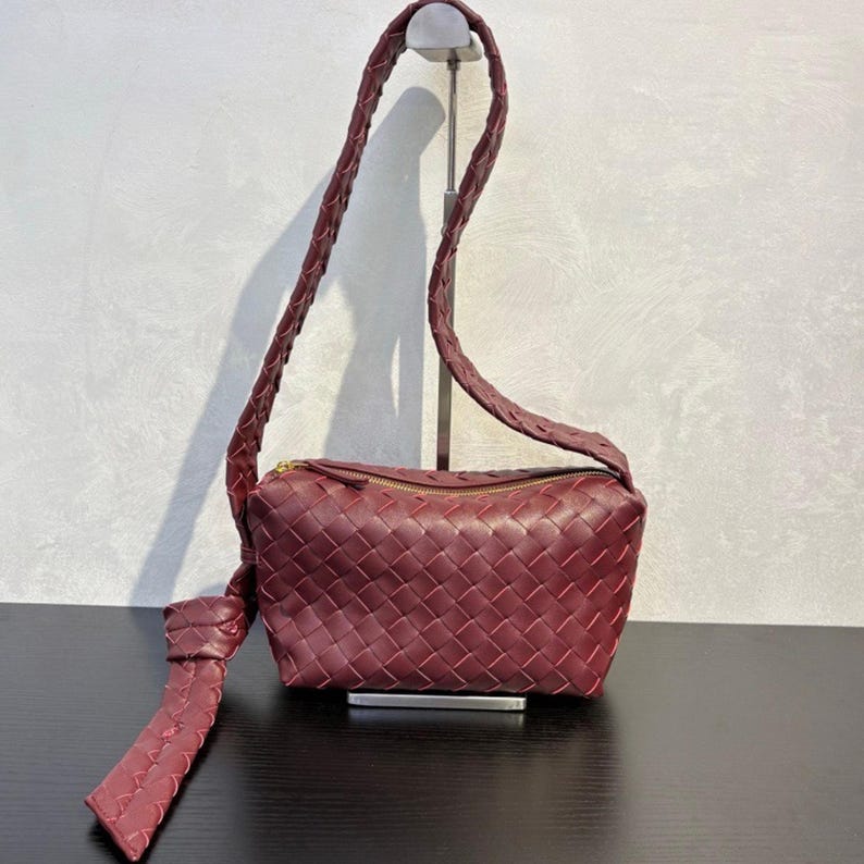 May include: A burgundy-colored crossbody bag with a woven design. The bag features a long, braided strap and a zippered top. A decorative knot detail hangs from the side. The bag is displayed on a black surface.