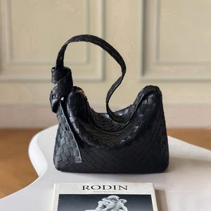 May include: Black woven leather shoulder bag with a knotted handle. The bag has a curved shape and a zipper closure. A book with the title "RODIN" is visible in the foreground.