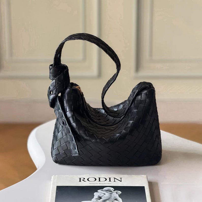 May include: Black woven leather shoulder bag with a knotted handle. The bag has a curved shape and a zipper closure. A book with the title "RODIN" is visible in the foreground.
