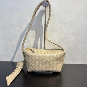 May include: Beige woven leather shoulder bag with a braided strap and a gold zipper. The bag has a rectangular shape and a long, braided strap. The bag is displayed on a black surface.