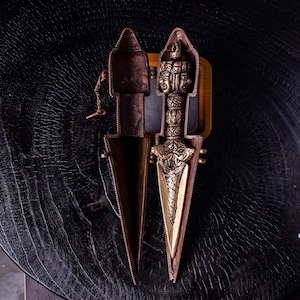 May include: A metallic phurba ritual dagger with an intricate three-faced deity handle and a triangular blade, resting inside its matching two-piece hinged metal case. The dagger features detailed engravings along the hilt and blade surface.