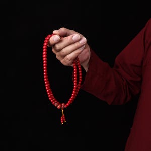 May include: A strand of small, round, vibrant red prayer beads featuring a smooth, polished finish. The beads are strung together on a thin cord, with a small yellow tassel hanging from the bottom, creating a circular loop of uniform red spheres.