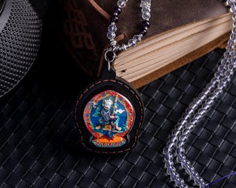 Puede incluir: Un collar con un colgante circular de madera negra que presenta una detallada pintura en miniatura de una deidad en tonos azul, rojo y dorado. La cadena está compuesta por cuentas transparentes facetadas y pequeñas cuentas esféricas moradas.