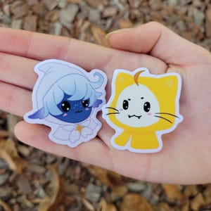 May include: Two cartoon character stickers. One is a blue and white character with a white hood and a star on its chest. The other is a yellow cat-like character with a white face and black eyes.