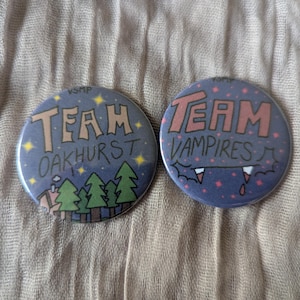 Puede incluir: Dos botones redondos. Uno dice "TEAM OAKHURST" con una escena de bosque, el otro "TEAM VAMPIRES" con colmillos. Ambos tienen un fondo azul con estrellas. Los botones están sobre una tela texturizada de tono neutro.