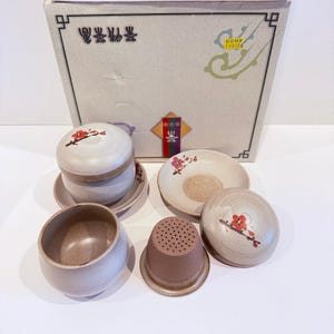 May include: A ceramic tea set with a beige and brown color scheme. The set includes a teapot, cups, saucers, and a strainer. Each piece is decorated with a red floral design. The set is presented in a cardboard box.