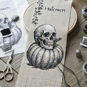 Könnte beinhalten: Ein Kreuzstichprojekt auf hellbeigem Stoff in einem hölzernen Stickrahmen, das einen schwarz-weißen Totenkopf auf einem Kürbis zeigt, mit dem Text Hallowen und einem Rankenmotiv, begleitet von Stickgarn und einer Stickschere.