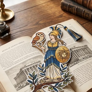 May include: A die-cut bookmark featuring a cross-stitch design of a figure in a gold helmet and blue dress holding a shield with a Medusa emblem and an owl. The bookmark includes a blue and gold tassel attached to the top corner of the design.