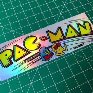 May include: A holographic sticker featuring the Pac-Man logo with the text "PAC-MAN by MIDWAY - Bally Co."