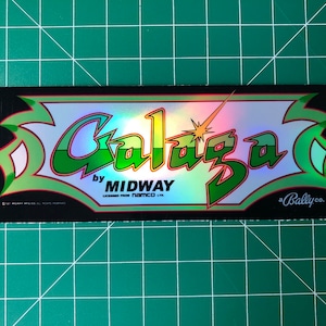 May include: A black and green arcade game marquee with the text "Galaga" in a rainbow iridescent font. The text "by Midway" is below the title in a smaller font. The text "Licensed from Namco Ltd." is below the text "by Midway". The text "a Bally Co." is in the bottom right corner of the marquee.