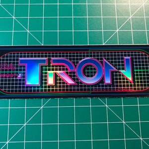 May include: A black and iridescent sticker with the word "TRON" in a grid pattern. The sticker also includes the text "Atari/Midway M. Productions".